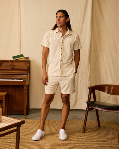 A man stands indoors wearing Dandy Del Mar's The Gaucho Terry Cloth Shorts in Alabaster with a matching short-sleeve shirt and white sneakers. A wooden piano, chair, and books set the scene for an Equatorial Cowboy vibe.