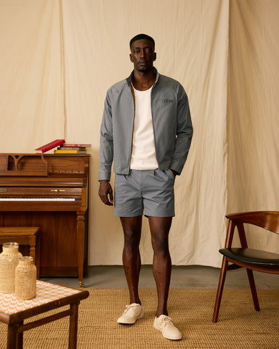 A man stands indoors wearing Dandy Del Mar’s The Rhodes Twill Shorts in Abyss, styled with a light blue jacket, white tank top, and beige shoes. In the background are a piano with books, a wooden chair, and a table.
