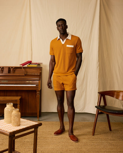 A man stands indoors wearing Dandy Del Mar’s The Cannes Waffle Knit Polo Shirt in Burnt Sienna, featuring a white collar and pocket detail. A wooden piano, chair, and table are visible in the background.