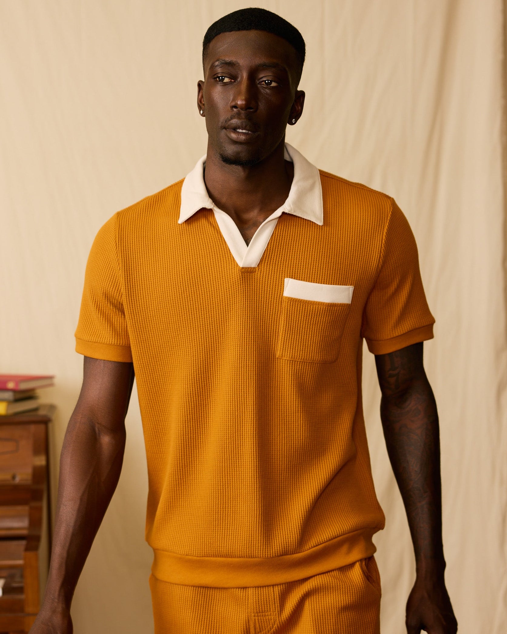 A man wears the Dandy Del Mar Cannes Waffle Knit Polo Shirt in Burnt Sienna, paired with matching shorts, standing by a brown piano topped with books against a cream backdrop.