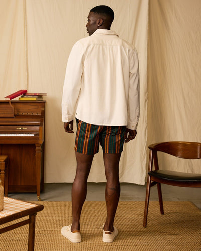 A man stands indoors facing away, wearing a cream shirt and Dandy Del Mar's The Mallorca Swim-Walk Shorts in Burnt Sienna with adjustable side fasteners. Slip-on shoes complete his look, with a wooden chair and piano in the background.