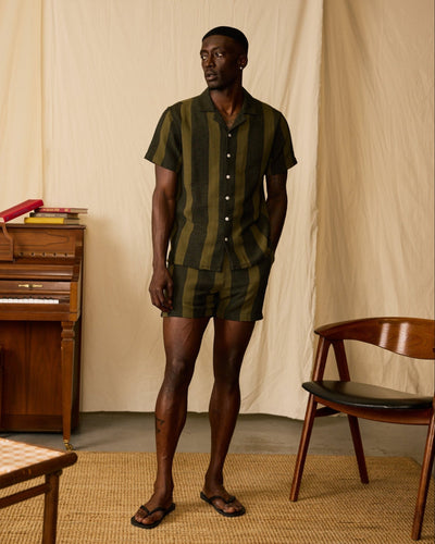 A man stands indoors by a wooden chair and piano, wearing Dandy Del Mar's The Palma Shorts - Onyx, a relaxed fit short in dark green and black yarn dyed stripe canvas, paired with a matching short-sleeved button-up shirt.