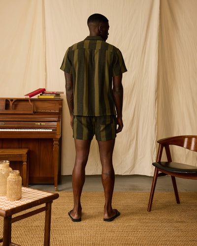 A man stands indoors with his back turned, wearing Dandy Del Mar's The Palma Shorts in Onyx, a short-sleeve green and brown yarn-dyed stripe canvas shirt, and black sandals. A piano, chair, and table appear in the background.