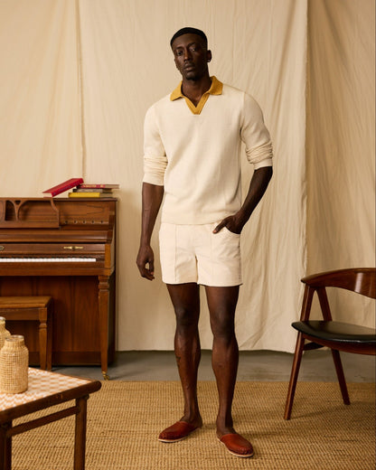 A man stands indoors in a cream knit sweater with a yellow collar, Dandy Del Mar’s The Corsica Corduroy Shorts - Scallop (5" inseam), and brown slip-on shoes. Behind him are a piano, books, chair, and table.