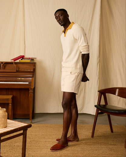A man stands indoors on a woven rug, wearing a cream sweater and The Corsica Corduroy Shorts - Scallop by Dandy Del Mar with red slippers. A wooden piano, stacked books, a chair, and a table are in the background.