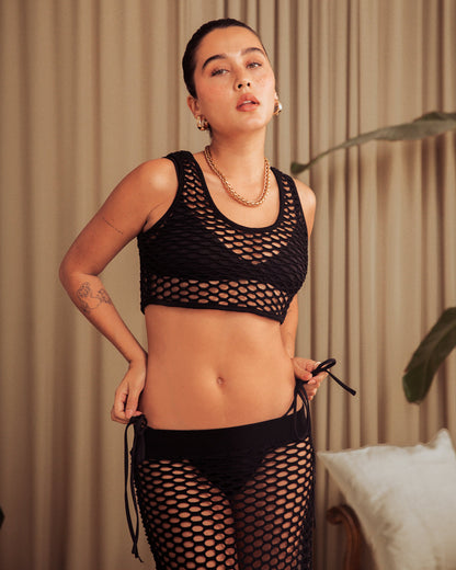 A person stands in front of a curtain, wearing The Montserrat Crochet Tank in Onyx from Dandy Del Mar and a matching bottom with cropped styling. Their hair is pulled back, and they have tattoos on one arm. They are surrounded by beige and green elements.