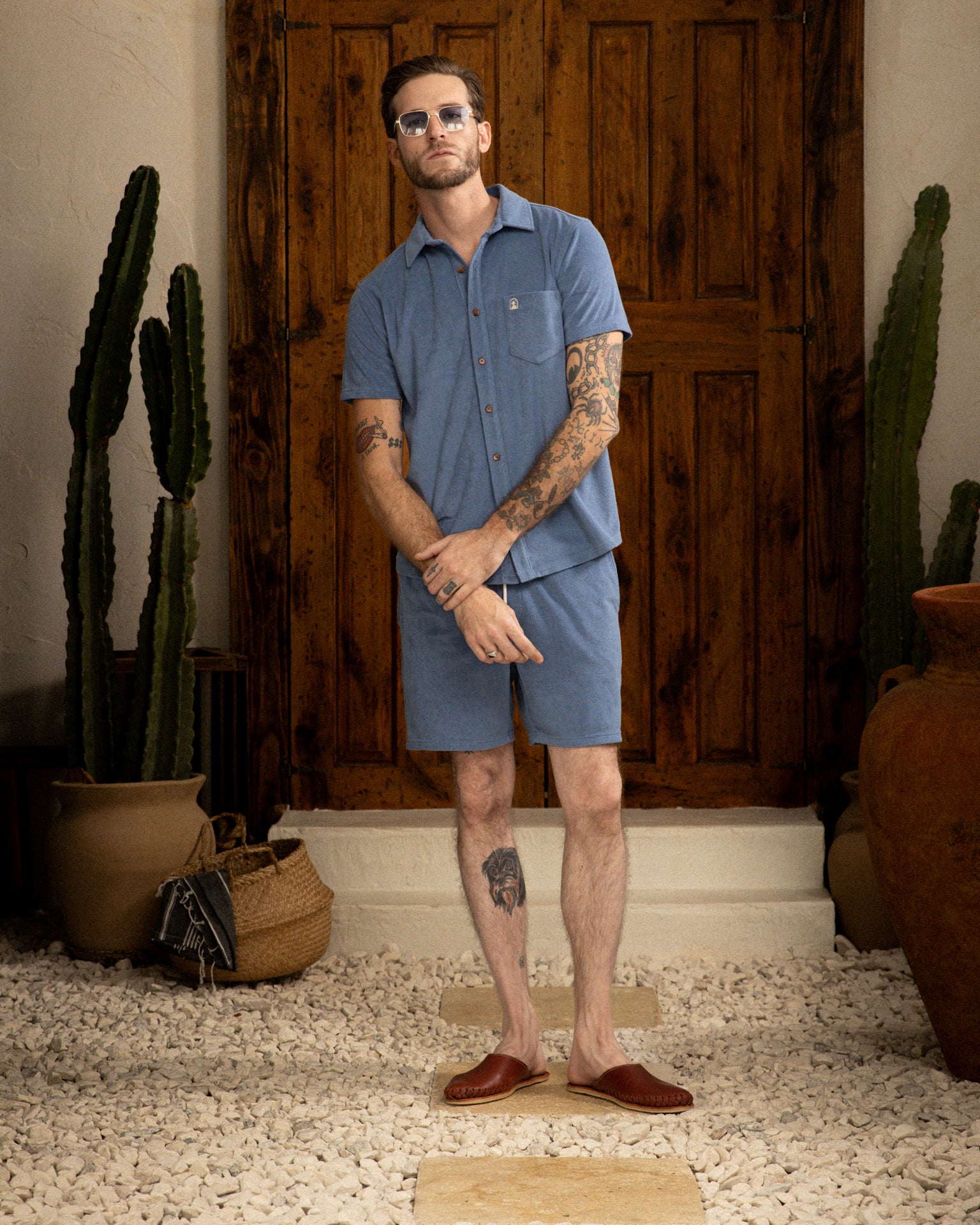 A man in a blue Annapolis Tropez Shirt and shorts from Dandy Del Mar, sporting sunglasses, stands with arms crossed before a wooden door. This premium terry cloth shirt embodies Côte d'Azur's relaxed luxury. Cacti and pots adorn the doorway.