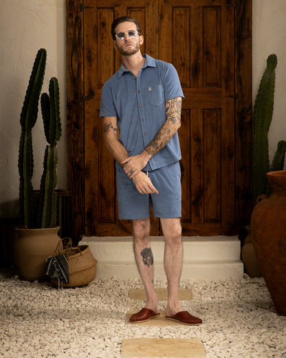 A man in a blue Annapolis Tropez Shirt and shorts from Dandy Del Mar, sporting sunglasses, stands with arms crossed before a wooden door. This premium terry cloth shirt embodies Côte d'Azur's relaxed luxury. Cacti and pots adorn the doorway.