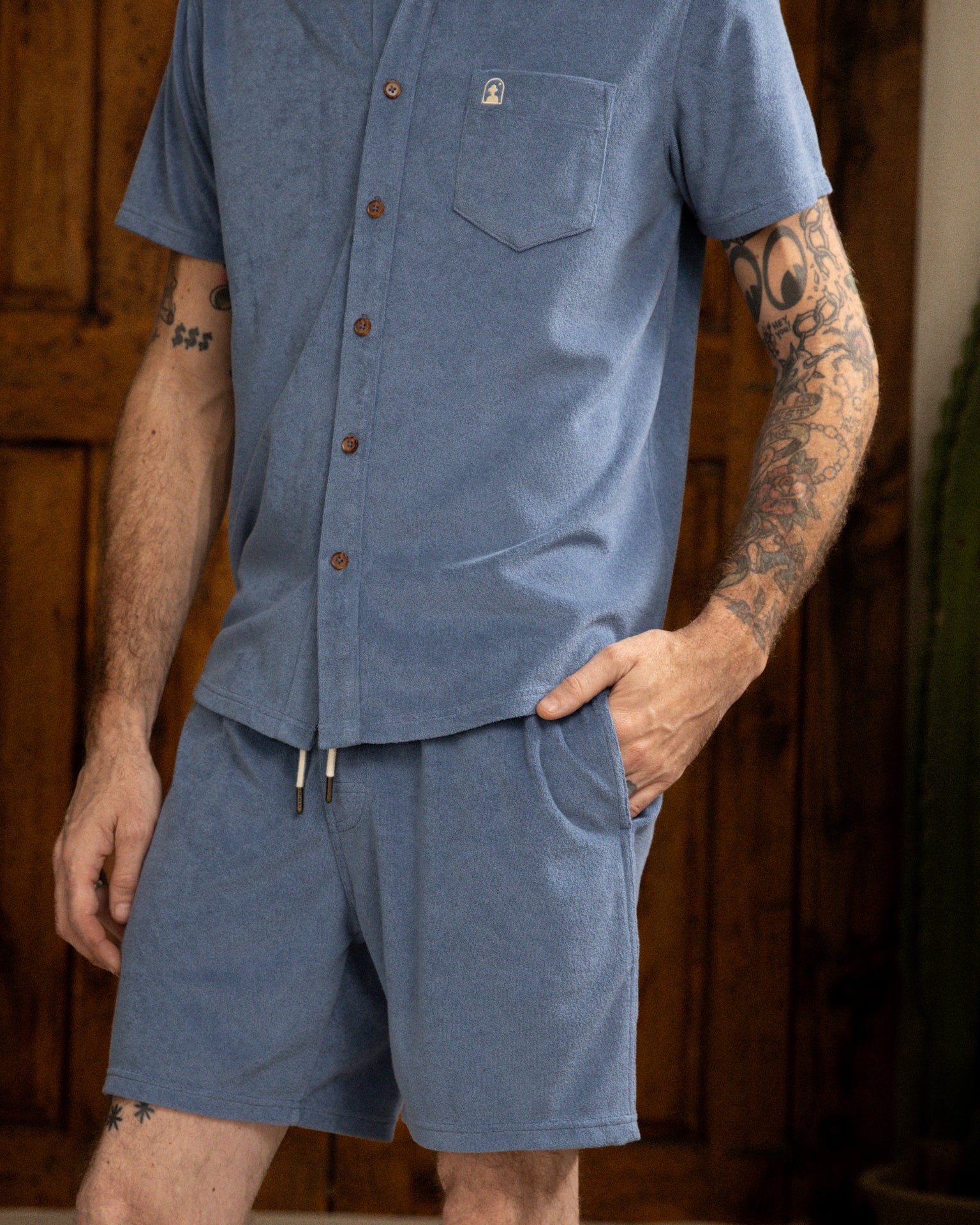 A person is sporting a blue button-up shirt and The Tropez Short - Annapolis by Dandy Del Mar, revealing their arm tattoos.