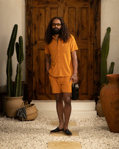 A person with long curly hair and glasses stands in front of a wooden door, wearing The Tropez Short in Burnt Sienna by Dandy Del Mar, capturing Côte d'Azur chic. They're holding a black book, with potted cacti on either side adding rustic elegance.