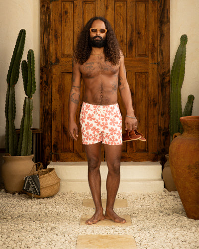 A man with long curly hair and sunglasses stands barefoot on stone, holding sandals and wearing Dandy Del Mar's Ventura Volley Shorts in Currant. Large cacti, pots, and a wooden door are in the background.