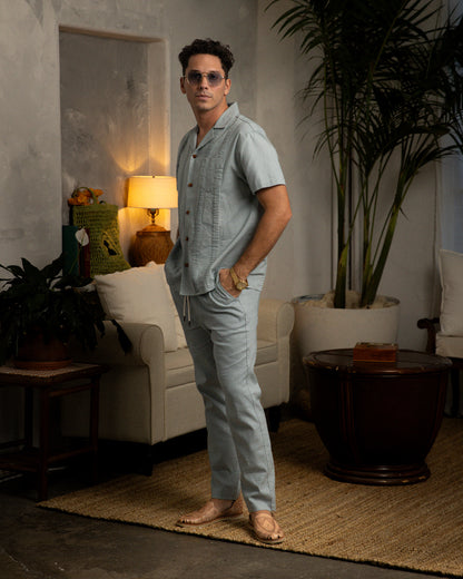 A man is standing in a living room wearing a Dandy Del Mar Brisa Linen Shirt featuring signature gardenia floral embroidery and coconut shell buttons.