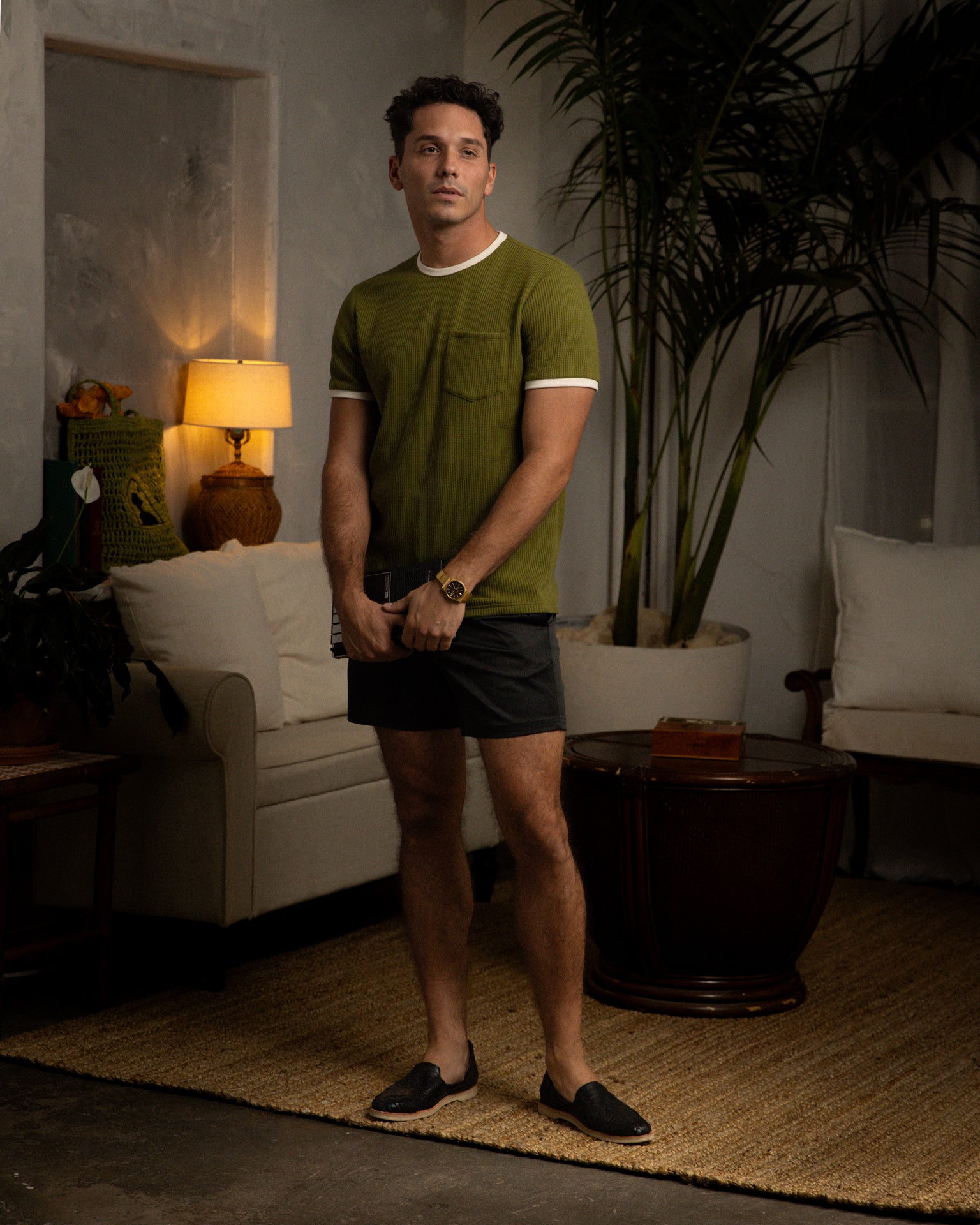 A man in The Cannes Waffle Knit Tee by Dandy Del Mar and dark shorts stands in a living room with a sofa, lamp, and plant. He holds a phone, wears a watch and slip-on shoes, exuding casual elegance.