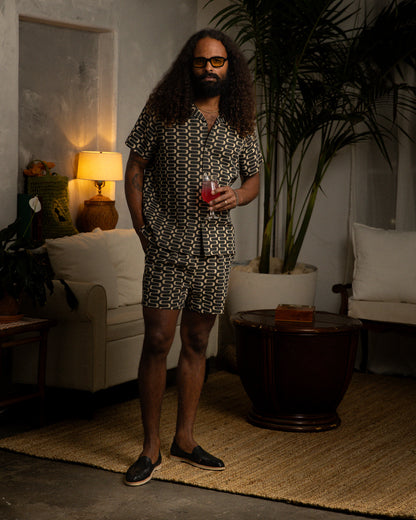 A lightweight man standing in a living room holding a drink while wearing a Dandy Del Mar Grenadine Shirt - Truffle.