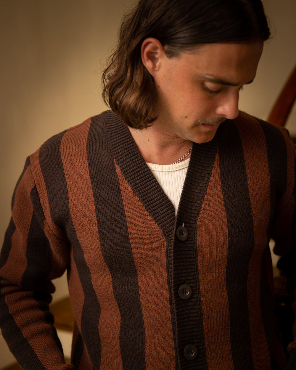 A person with long hair wears The Florence Knit Cardigan - Garam by Dandy Del Mar, featuring a brown and black jacquard knit pattern and ribbed trims, over a white shirt, looking down.