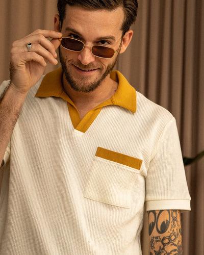 A bearded, tattooed man lifts his sunglasses and smiles, wearing the Dandy Del Mar Cannes Waffle Knit Polo Shirt in Vintage Ivory with mustard trim. A curtain is in the background.
