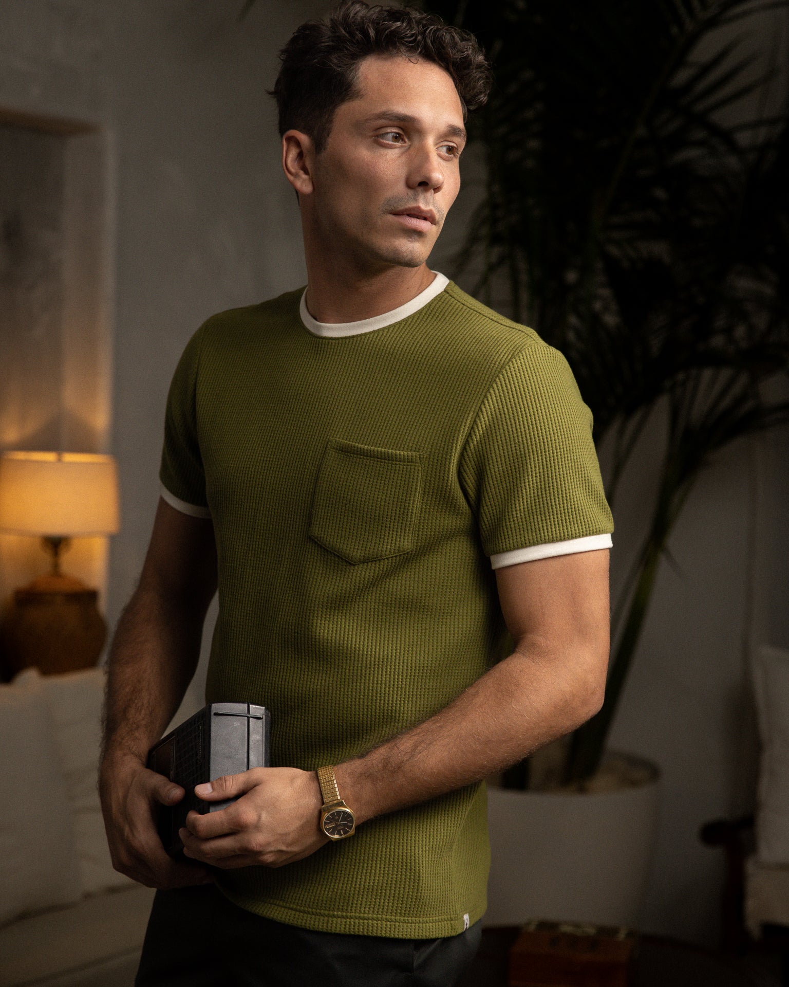 A person wearing The Cannes Waffle Knit Tee in Arbequina by Dandy Del Mar holds a device indoors, with a lamp and plants visible, creating a cozy atmosphere.