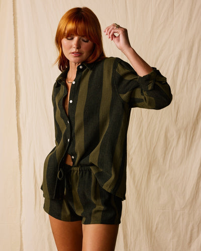 A person in the Dandy Del Mar Sevilla Shorts - Onyx, featuring a matching olive and black striped button-up and elastic waistband, stands before a beige fabric backdrop.