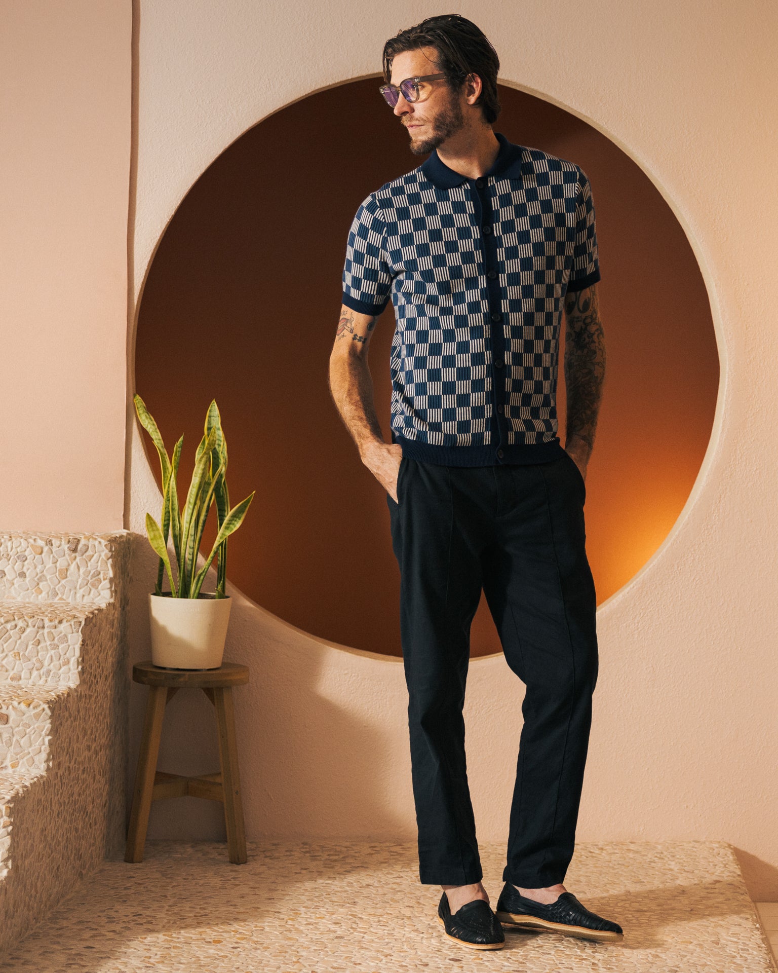 A bearded man wearing glasses, The Martinique Sweater-Knit Shirt in Anchor by Dandy Del Mar, black pants, and black shoes stands near a plant on a stool, with a circular orange backdrop behind him.