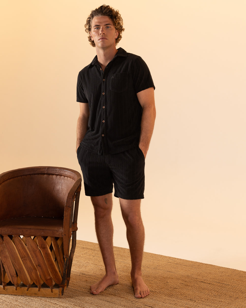 A person stands barefoot by a wooden chair on a woven rug, wearing Dandy Del Mar's The Tropez Terry Cloth Short in Onyx and a black short-sleeve button-up shirt, against a beige background.