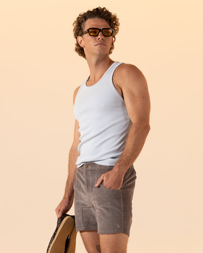A man wearing The Corsica Corduroy Shorts in Argento by Dandy Del Mar, a white tank top, and sunglasses stands holding a hat, looking off to the side against a light beige background.