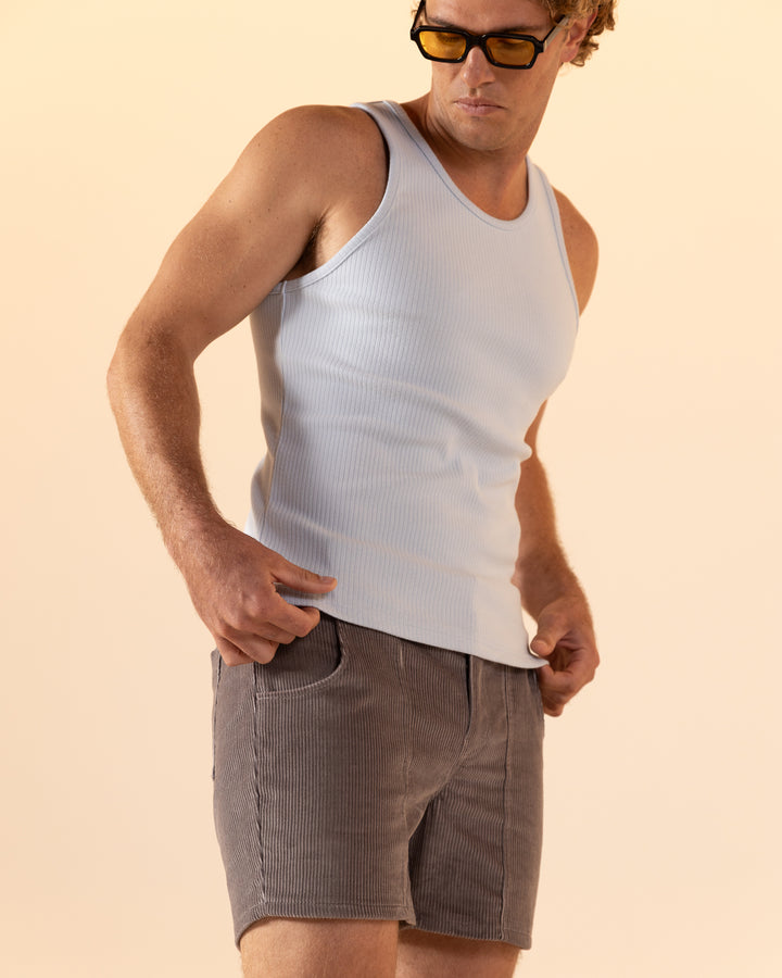 A man stands with hands in pockets against a neutral background, wearing The Milan Rib Tank in Comet Grey by Dandy Del Mar and gray shorts, paired with sunglasses.