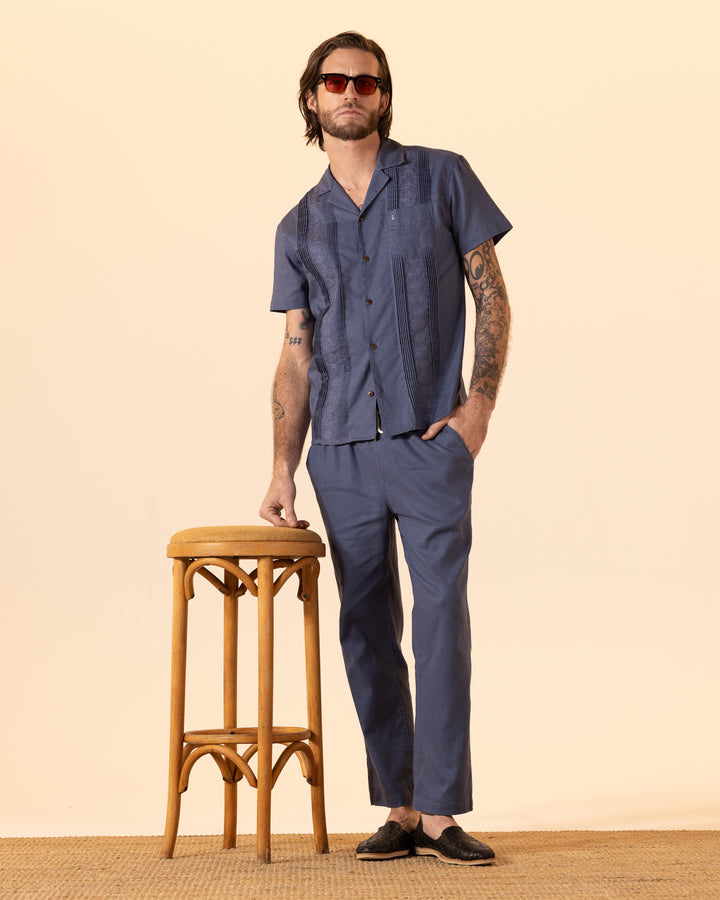 A man in Dandy Del Mar's blue Brisa Linen Shirt - Moontide and pants leans on a stool. He sports sunglasses and black loafers against a beige background.