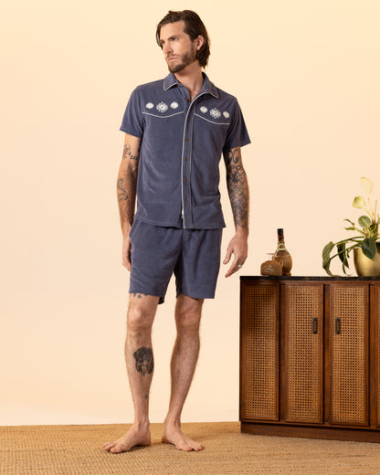 A man stands barefoot beside a wooden cabinet with plants and a bottle, wearing Dandy Del Mar's The Gaucho Short - Moontide set, featuring a blue short-sleeve shirt with a floral design and matching gaucho shorts with an elastic waistband.