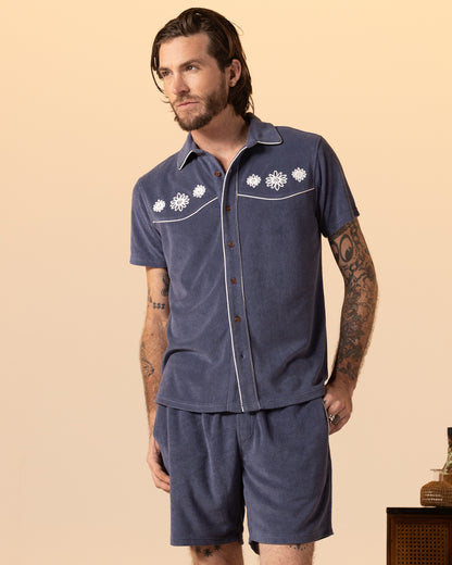 Tattooed man wearing Dandy Del Mar's The Gaucho Short in Moontide, a blue short-sleeve terry cloth shirt with matching elastic waistband shorts, stands against a beige background.