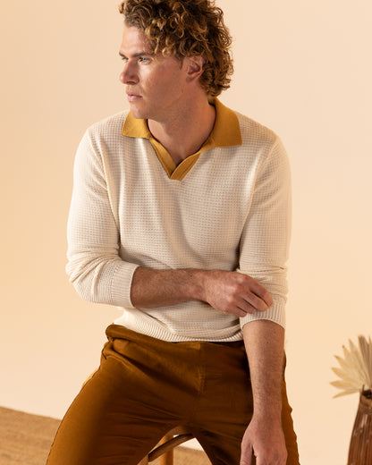 Seated on a stool, an individual in Dandy Del Mar's The Sebastian Long Sleeve Polo in Vintage Ivory with matching pants gazes sideways. The textured ensemble blends seamlessly with the neutral beige background.