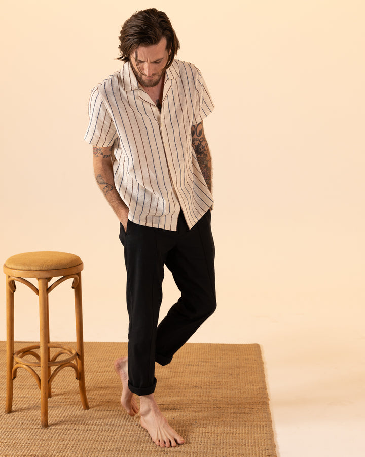 A tattooed person, barefoot on a woven mat and wearing Dandy Del Mar's "The Quarta Shirt - Scallop Weave" with black pants, stands next to a wooden stool.