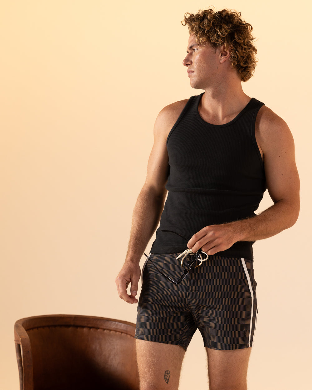 Someone wearing the Onyx Stirata Swim Short from Dandy Del Mar stands near a brown chair, looking to the side against a beige backdrop.