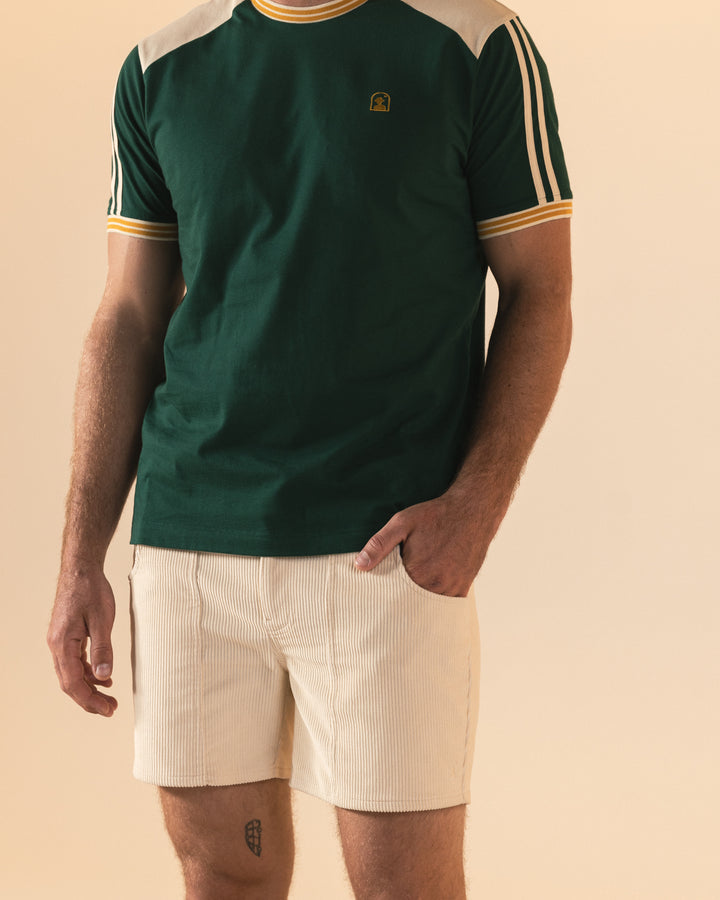 A man in a Dandy Del Mar Nassau Tee - Albero in green and white, with striped shoulders and an embroidered logo, stands casually with one hand in his pocket, paired with beige shorts.