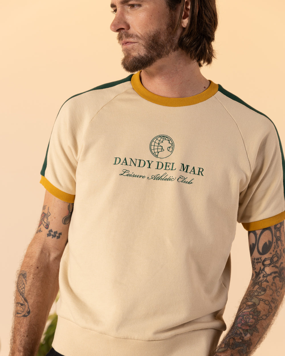 A tattooed man with a beard is wearing The Marseille French Terry Pullover - Sabbia by Dandy Del Mar, a cream-colored shirt with green and yellow trim featuring the text "Dandy Del Mar Leisure Athletic Club.