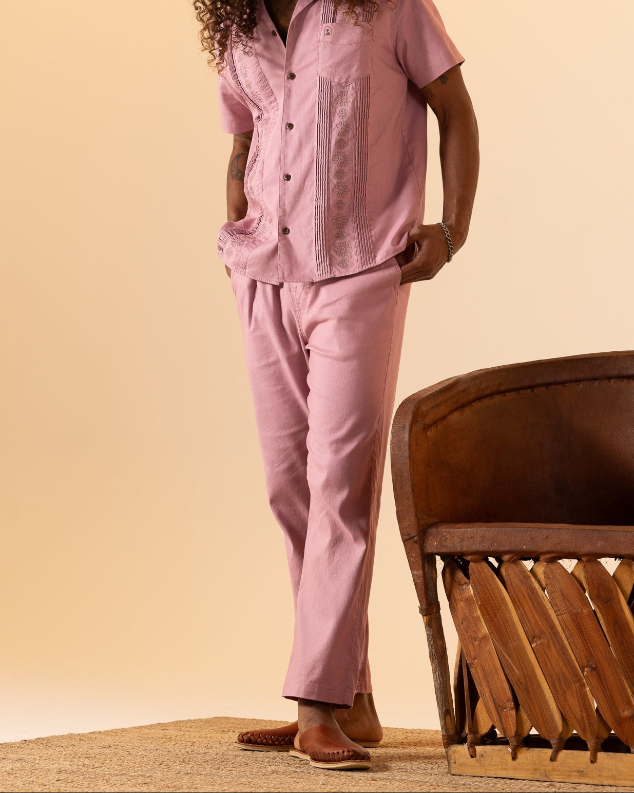 A person with long curly hair wears a pink short-sleeve shirt and The Brisa Linen Pant - Dawn by Dandy Del Mar, standing near a brown chair on a carpeted floor.
