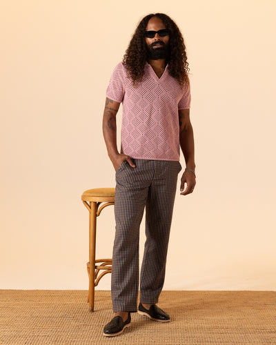 A man with long curly hair and a beard stands indoors wearing sunglasses, plaid trousers, loafers, and the Dandy Del Mar Antibes Crochet Polo Shirt in Dawn, next to a wooden stool.