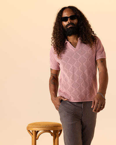 A man with long curly hair and sunglasses stands by a wooden stool, wearing Dandy Del Mar’s The Antibes Crochet Polo Shirt in Dawn and plaid pants against a beige background.