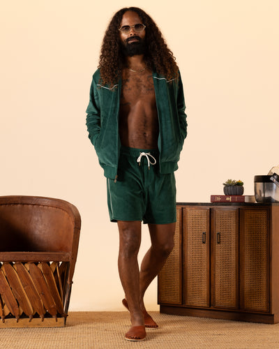 A man with long curly hair and glasses wears a green velour jacket and Dandy Del Mar's The Rio Velour Shorts - Albero, featuring an elastic waistband, while standing barefoot in a living room beside a wooden cabinet and chair.