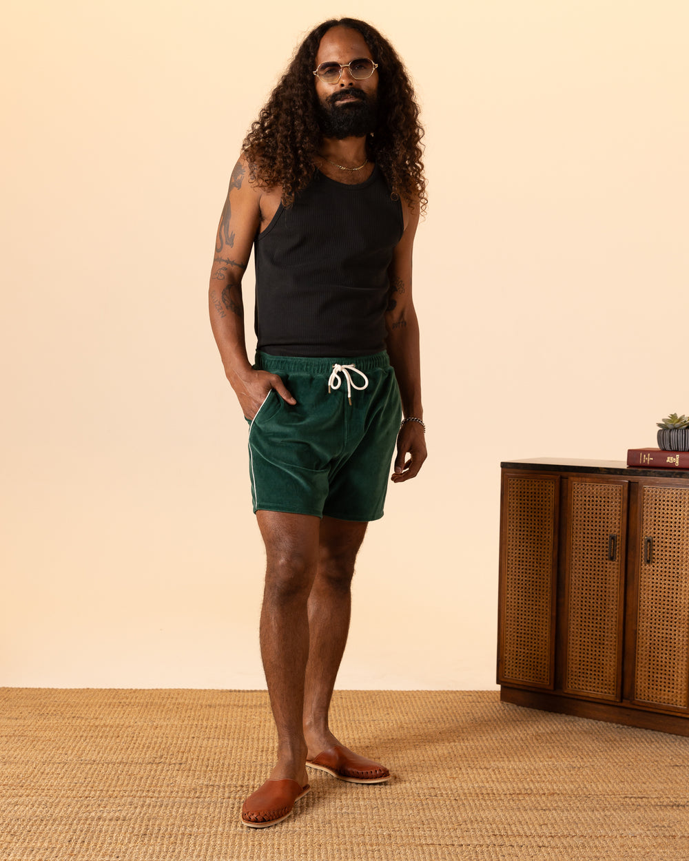 A man with long curly hair and tattoos stands wearing a black tank top, The Rio Velour Short - Albero by Dandy Del Mar, and sandals in front of a wooden sideboard.