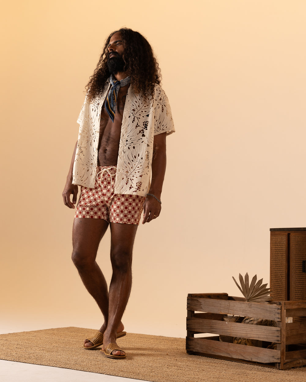 A man with long hair in a white openwork shirt and sandals stands on a woven mat beside a wooden crate against a light background, wearing Dandy Del Mar’s The Ventura Volley Short in Rouge.