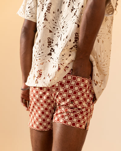 A person wearing a cream crochet short-sleeve top and Dandy Del Mar's The Ventura Volley Shorts - Rouge, featuring a red-and-white geometric pattern, elastic waist, and one hand tucked in the pocket.