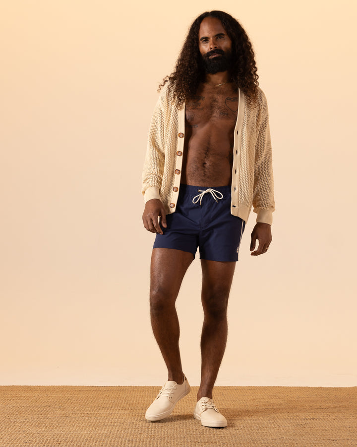 A person with long curly hair wears a beige cardigan, The Ventura Volley Short in Anchor by Dandy Del Mar, and beige shoes, standing on a woven rug against a neutral background.