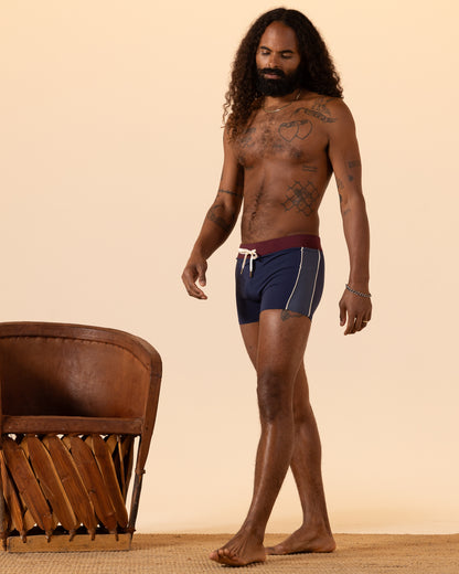 A man with long hair and tattoos stands beside a brown chair on a rug, wearing The Cassis Square Cut Swim Brief - Anchor Bordeaux by Dandy Del Mar.