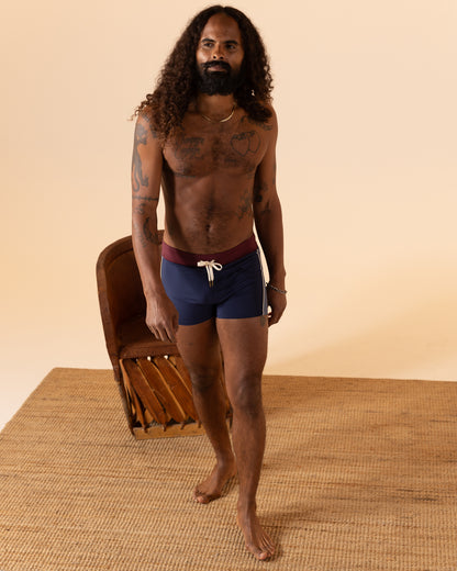 A person with long curly hair stands on a woven mat near a brown chair, wearing Dandy Del Mar's Anchor Bordeaux Cassis Square Cut Swim Brief, featuring a maroon waistband and white drawstring, made from recycled polyester.