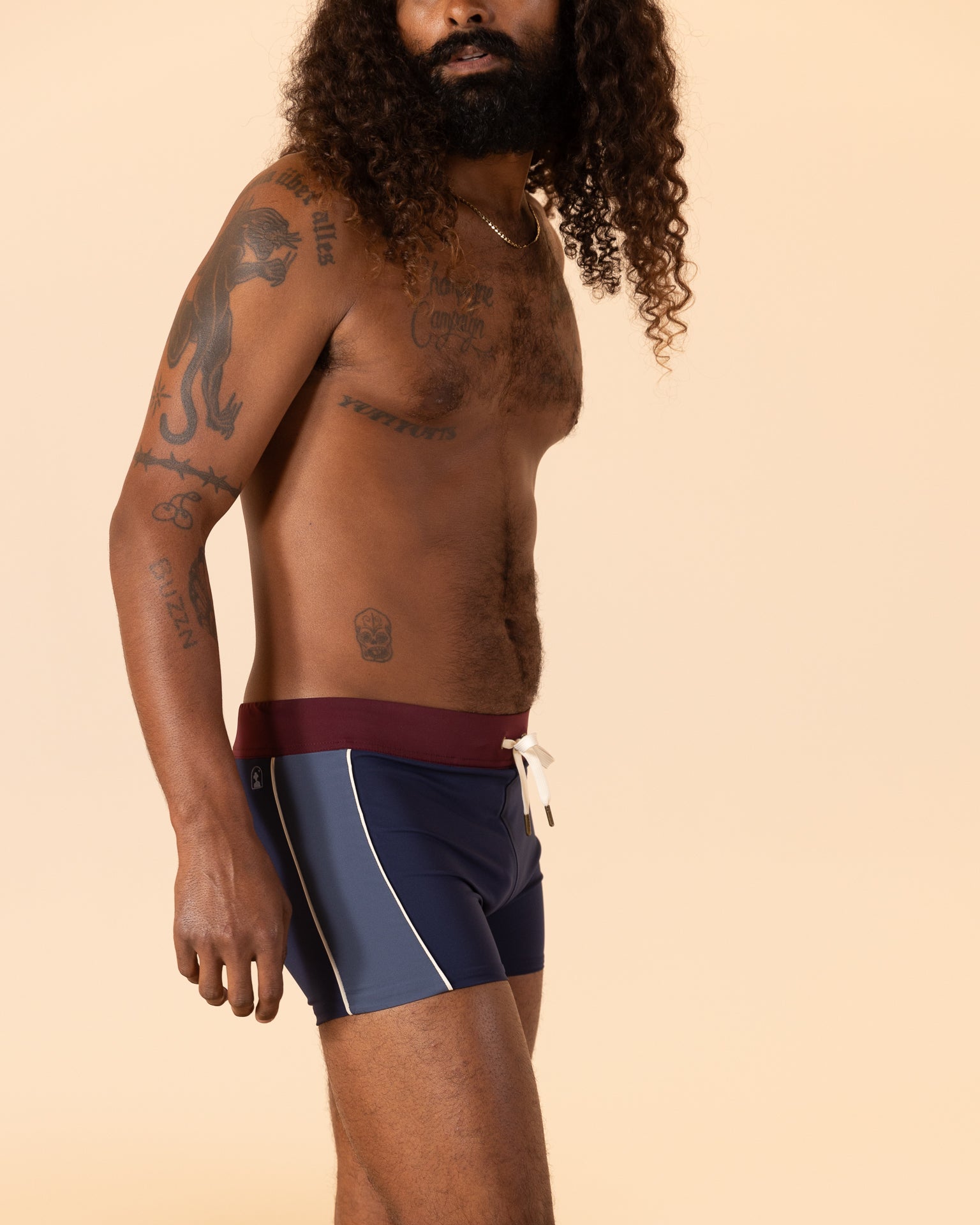 A man with long curly hair stands in profile, highlighting his European fit wearing The Cassis Square Cut Swim Brief - Anchor Bordeaux by Dandy Del Mar. Made from recycled polyester, the swimwear complements his intricate tattoos against a plain beige backdrop.