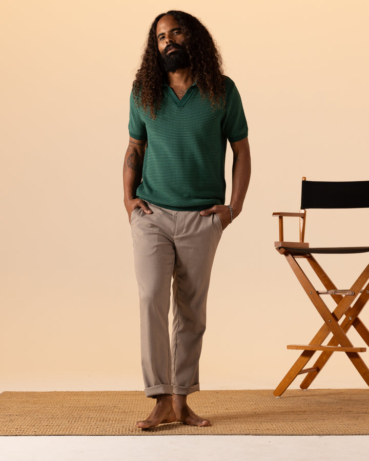 A person with long curly hair, barefoot on a mat, stands next to a director's chair in a plain setting. They are wearing beige pants and The Palamos Knit Polo Shirt - Albero by Dandy Del Mar.