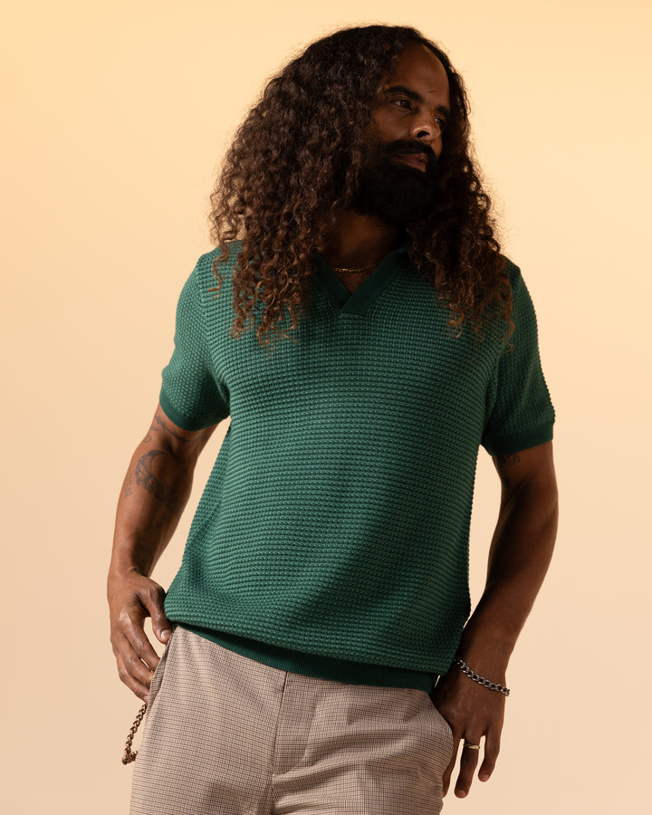 A bearded individual with long curly hair wears The Palamos Knit Polo Shirt - Albero by Dandy Del Mar, paired with light-colored pants, standing against a plain background.