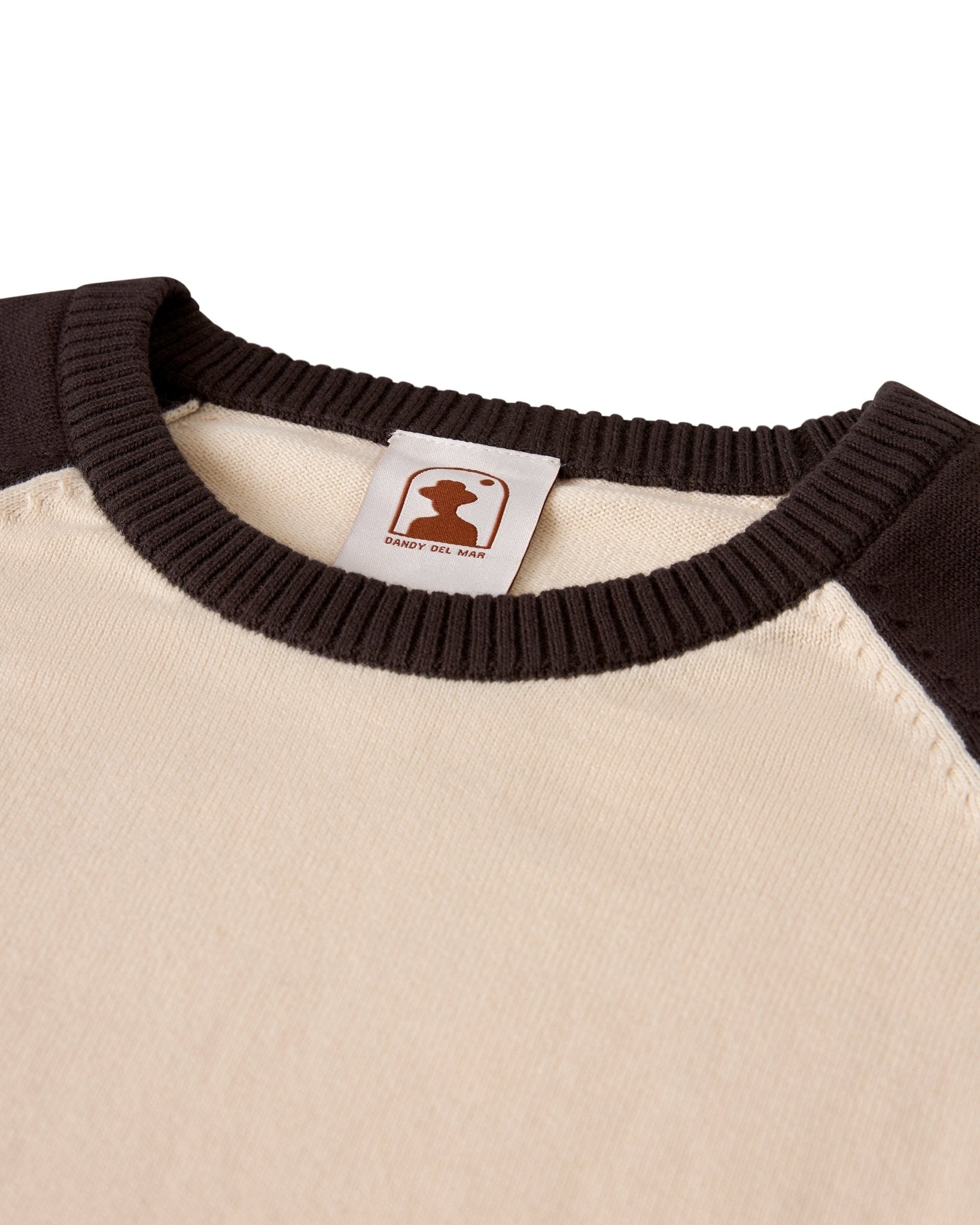 Close-up of The Grenada Sweater Knit Baseball Tee - Tierra by Dandy Del Mar—a beige and brown lightweight cotton crewneck with ribbed collar, featuring a silhouette logo and "SABOR DEL MAR" label for vintage-sport style.