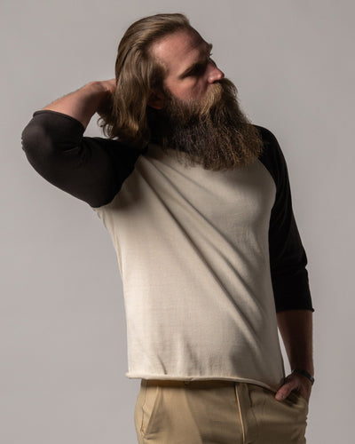 A bearded man stands sideways, one hand behind his head, wearing Dandy Del Mar's The Grenada Knit Baseball Tee - Tierra in cream and black with beige pants.