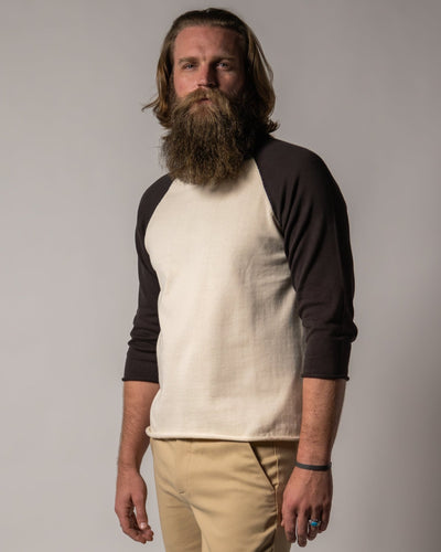 A man with long hair and a beard wears the Dandy Del Mar Grenada Knit Baseball Tee - Tierra, featuring dark sleeves and lightweight cotton, paired with beige pants, standing against a plain backdrop for a vintage-sport vibe.